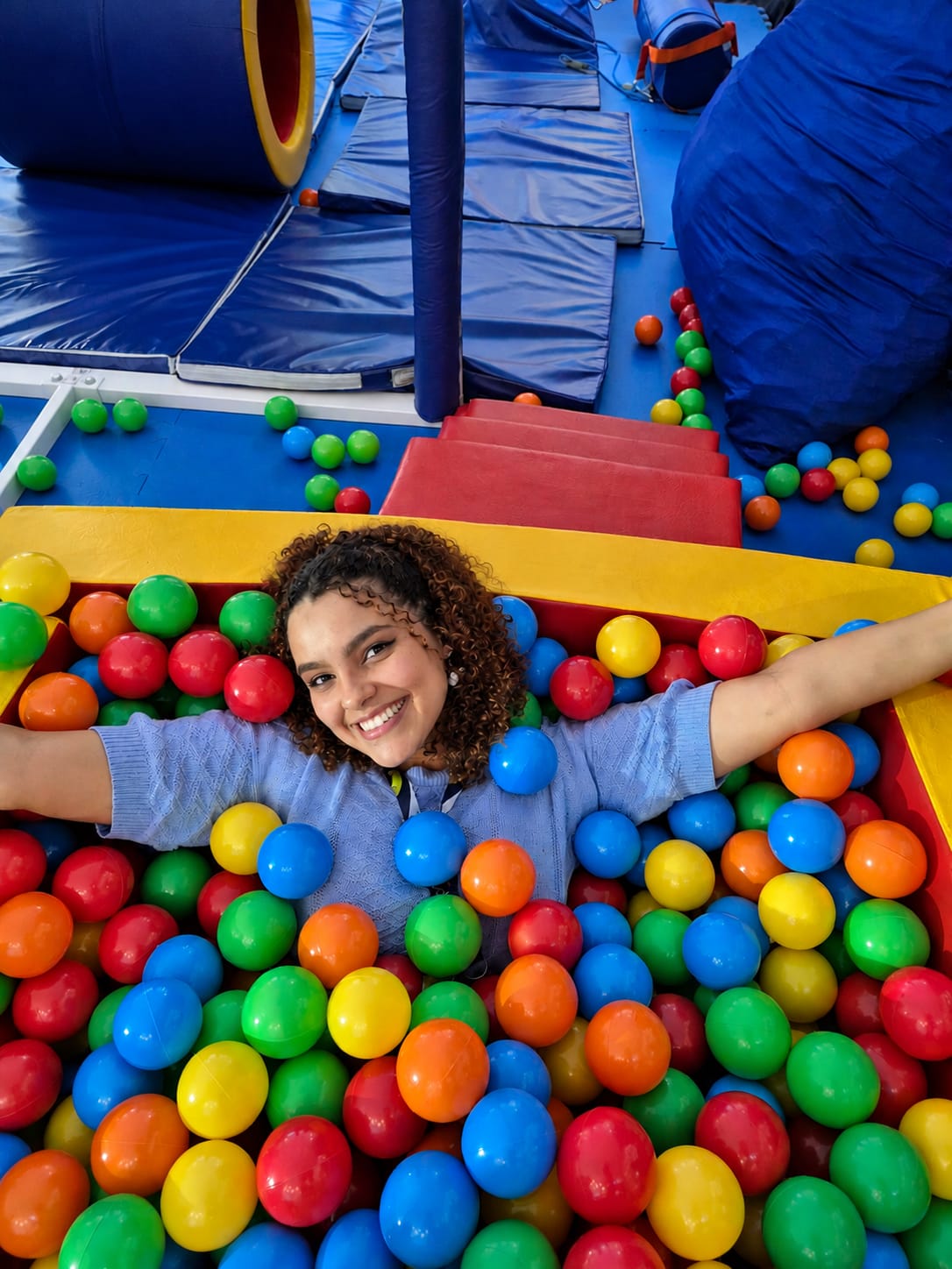 Júlia Patrocínio Aguiar, terapeuta ocupacional, sorrindo de braços abertos dentro de uma piscina de bolinhas coloridas